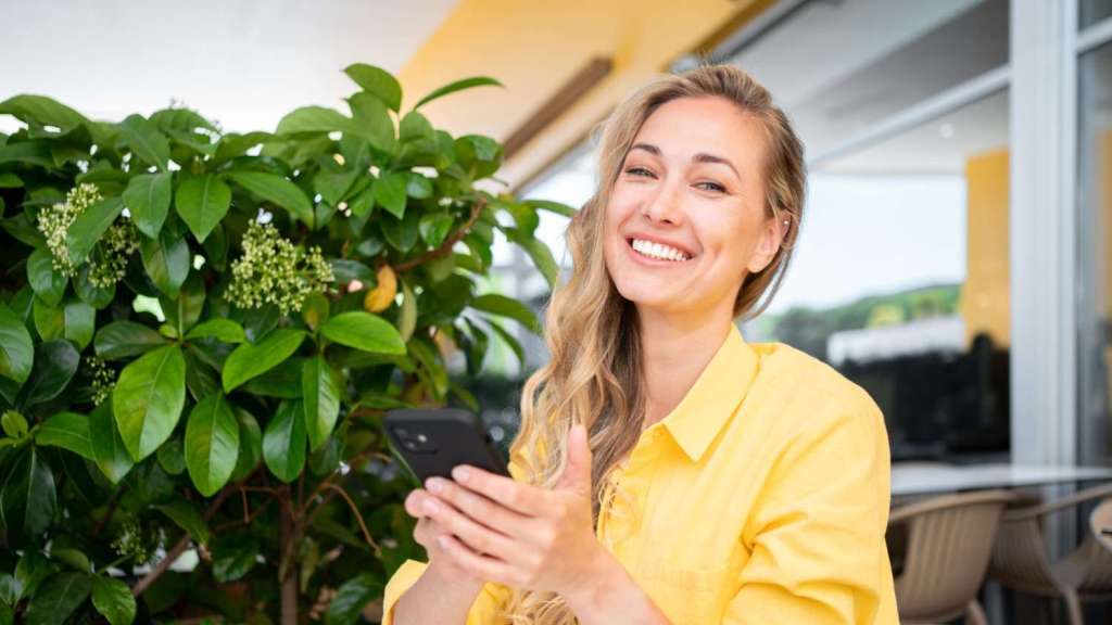 A young female with long blonde hair wears a yellow shirt and smiles as she sits outside next to a bush and holds her phone, reading an article about how to become sole trader in the UK