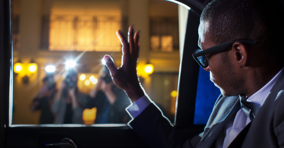 A man in a car waves to people outside as people take photographs