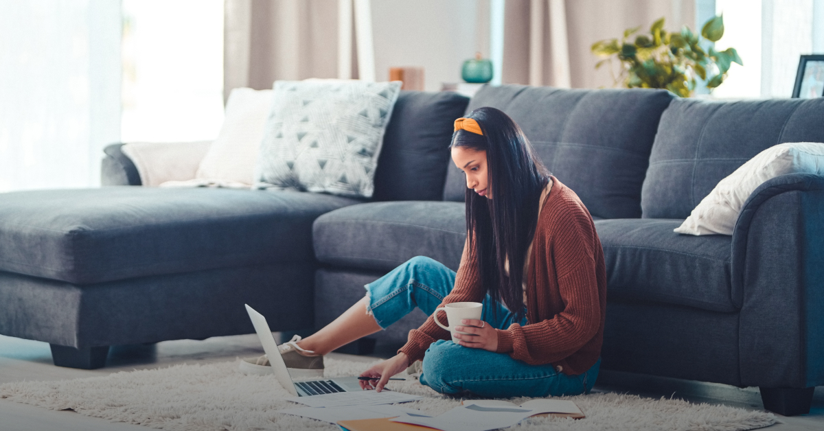 A girl with dark hair sits on the floor in front of her sofa, reading an article on her laptop about the cost of living in the UK