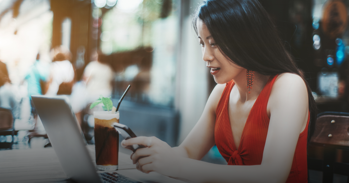 A young woman sits in a cafe with a drink and her laptop on the table while reading about buying a property in the Philippines on her mobile phone