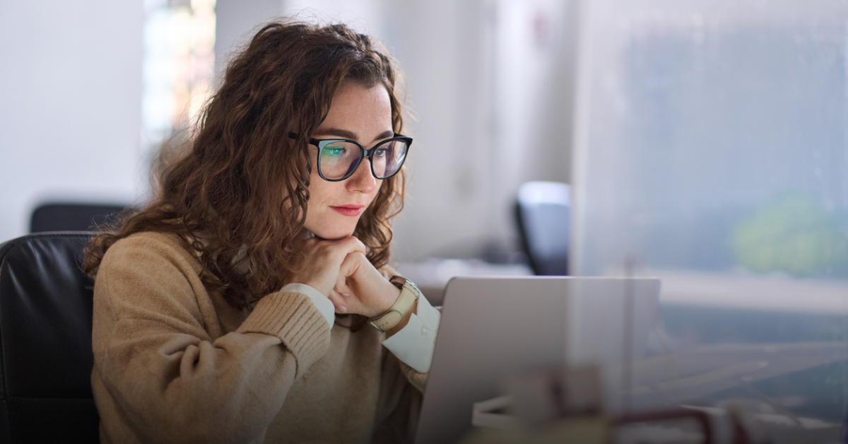 A young woman with brown curly hair and glasses sits at a table and reads an article about the best Revolut alternatives on her laptop