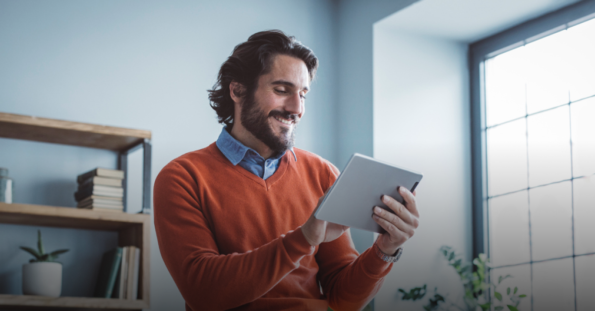 A man with brown hair wears a blue shirt and orange jumper and smiles while looking at his tablet reading an article about Wise vs OFX features