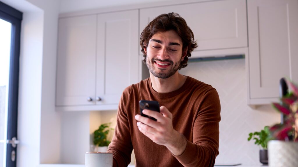 A young man smiles to himself as he stands in his kitchen reading the TransferGo vs Revolut Business comparison article on his mobile phone