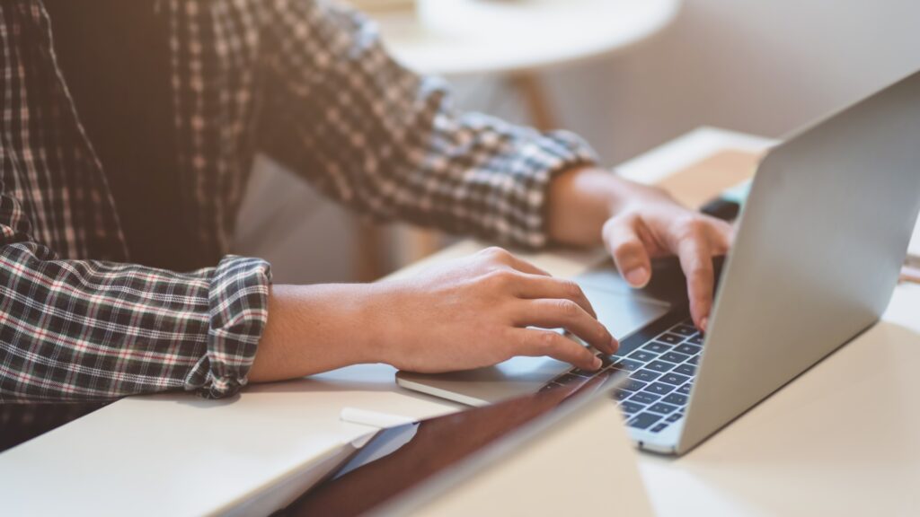 A man in a checked shirt sits at a table reading an article on his laptop about Lloyds Business Accounts.