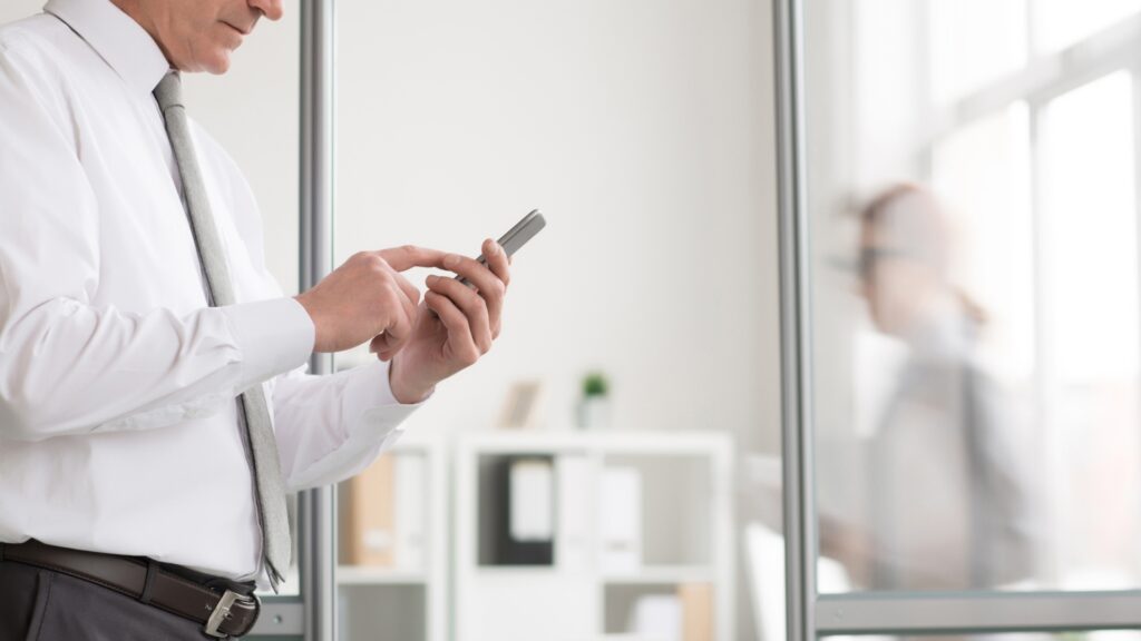 A man in a white shirt stands in his office, reading an article on his mobile phone about 'What are b2b payments?'