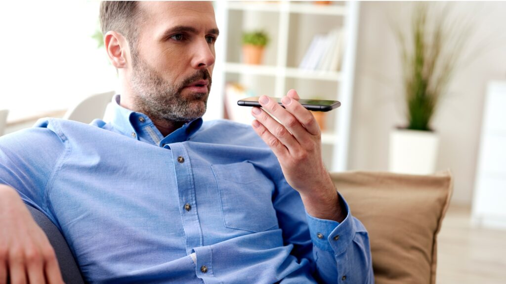 A man in a blue shirt sits on his sofa and talks to his smartphone on speaker to his Lloyds Business Account contact.