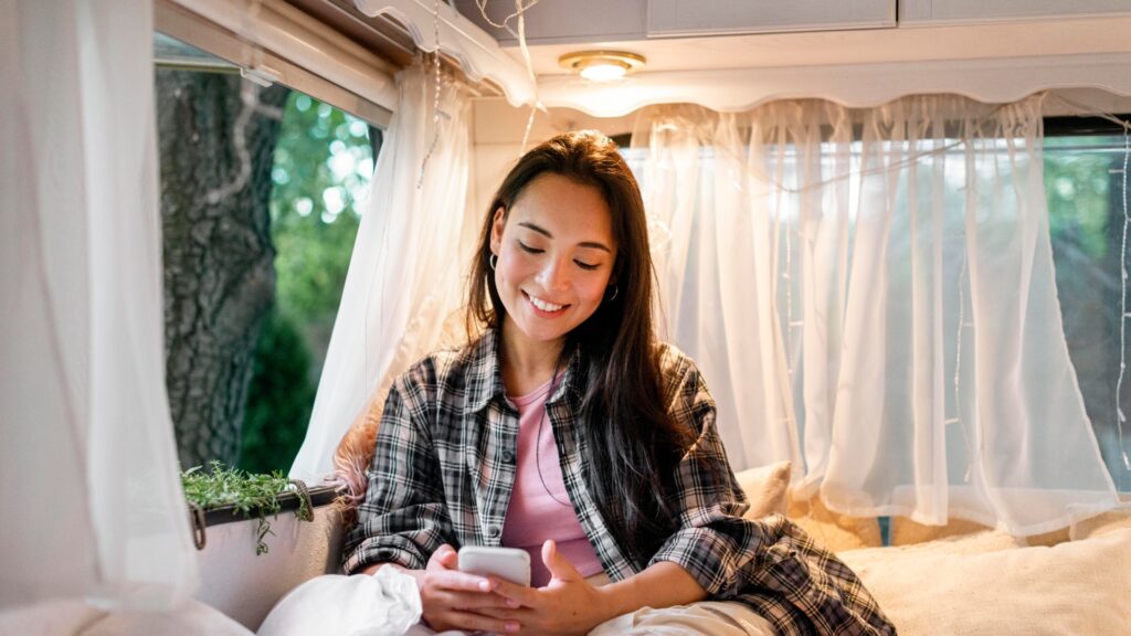 A relaxed and smiling young female sits in a campervan reading an article on her smartphone on the topic of, 'Is Lloyds Business safe?'