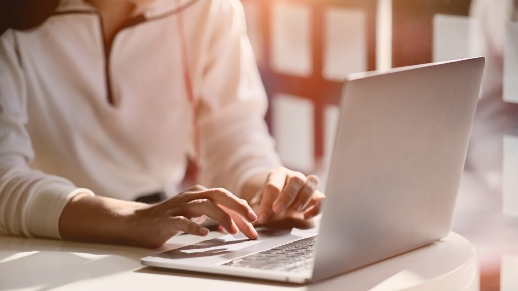 A person in a white sweatshirt sits at a table and types on their laptop while considering a Lloyds Business Bank Account.