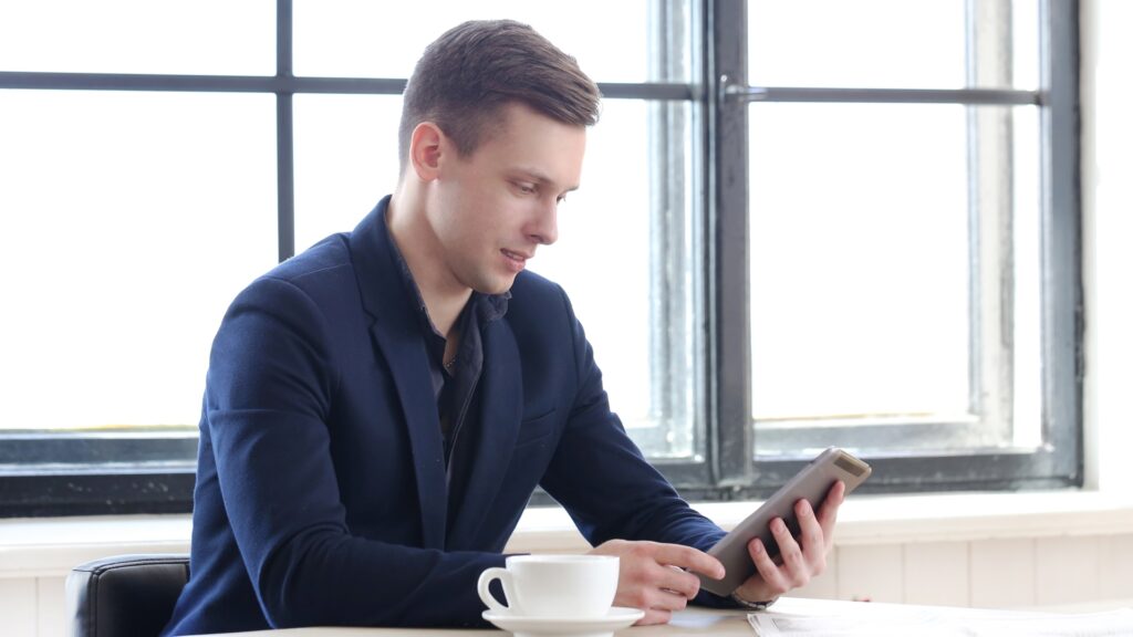 A young white male sits at his desk and reads an article on his tablet titled, 'Is Lloyds Business safe?'