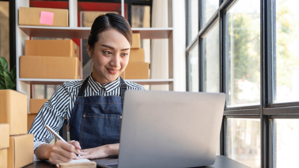 A young brunette female in a uniform sits at her desk and writes as she looks at her laptop to read an article about how to open business bank account in the UK