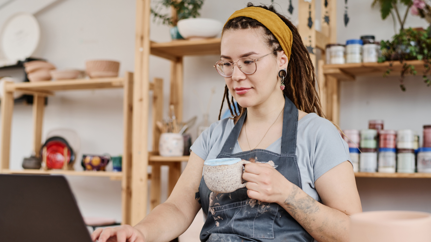 A young brunette female stands in a cafe, holding a coffee and typing on her laptop as she researches how to open business bank account in the UK