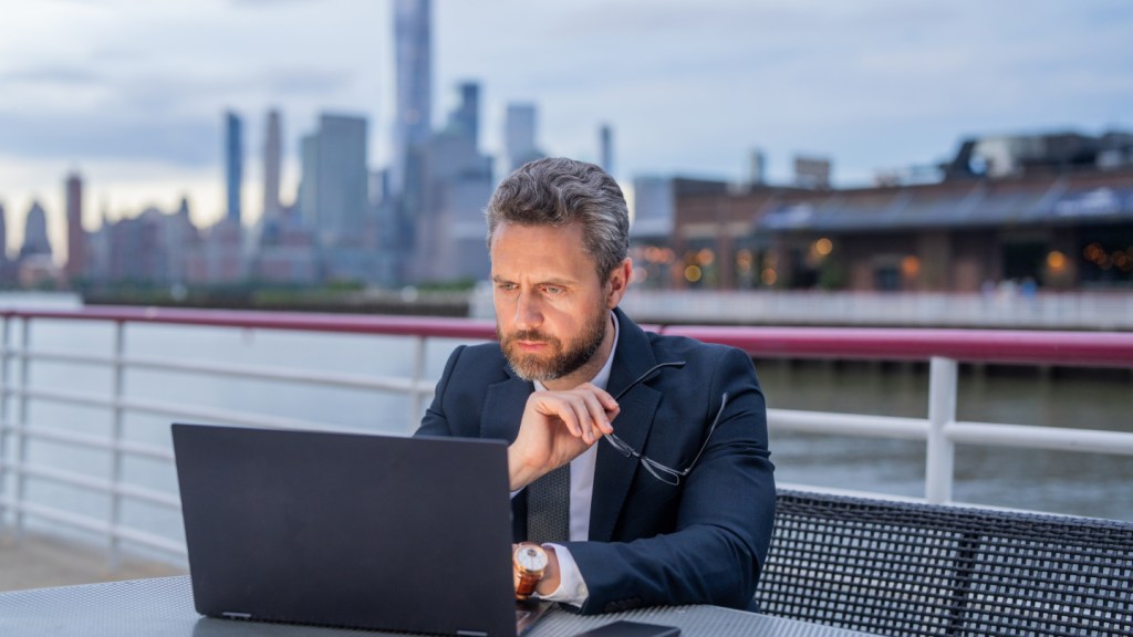 A middle-aged man with a city backdrop behind him sits on a bench outdoors by the river, holding his glasses and reading an article on his laptop about how to open business bank account in the UK