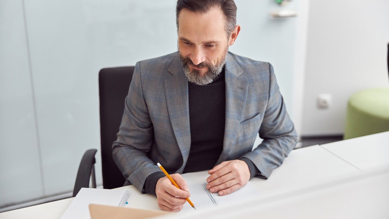 A bearded man in a grey suit sits at a desk and writes with a pen and paper as he commences the process to open bank account in Lithuania