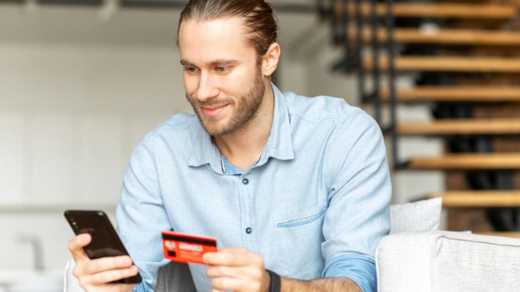 A young bearded male in a blue shirt sits at a desk with his mobile and his banking card as part of his process to open bank account in Lithuania