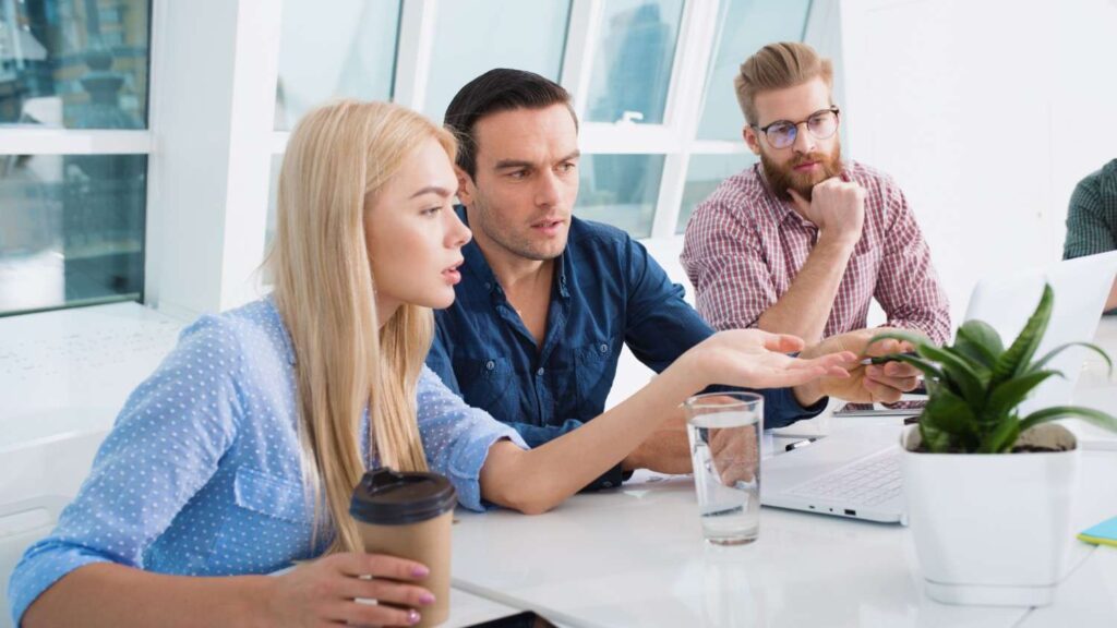 A young blonde female sits at a table drinking coffee next to two young men. Together, they read an article on a laptop about Lithuanian business culture