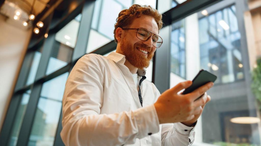 A bearded man wearing glasses and a white shirt smiles to himself as he checks his phone to ask the question 'Is Revolut Business safe?'