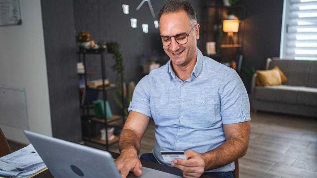 A man in a pale blue shirt and glasses sits at a table in his living room, whilst looking at his bank card and laptop and asking the question 'Is Revolut Business safe?'