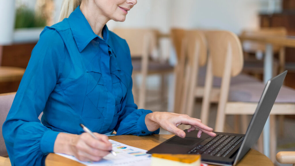 A young blonde female in a blue shirt sits at a table and simultaneously writes and types on her laptop to prepare for her Lithuania Startup Visa application