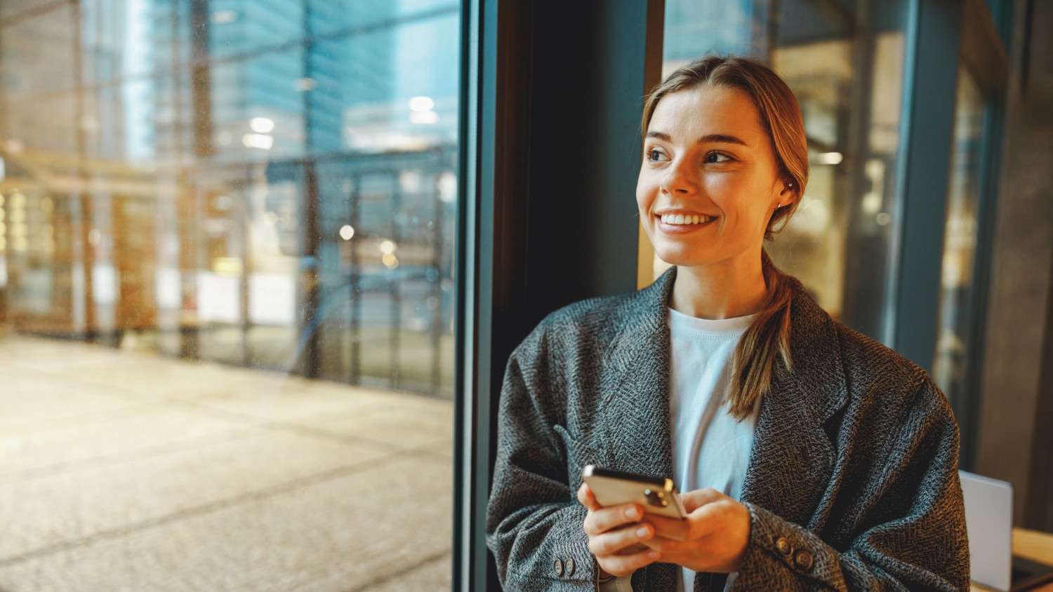 A young woman smiles as she looks out of the window while holding her phone and reading an article about TransferGo vs Luminor Business