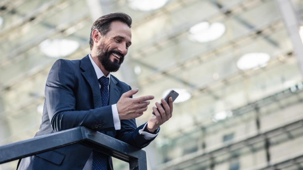 A bearded male in a suit smiles as he reads an article on his phone about TransferGo vs Luminor Business