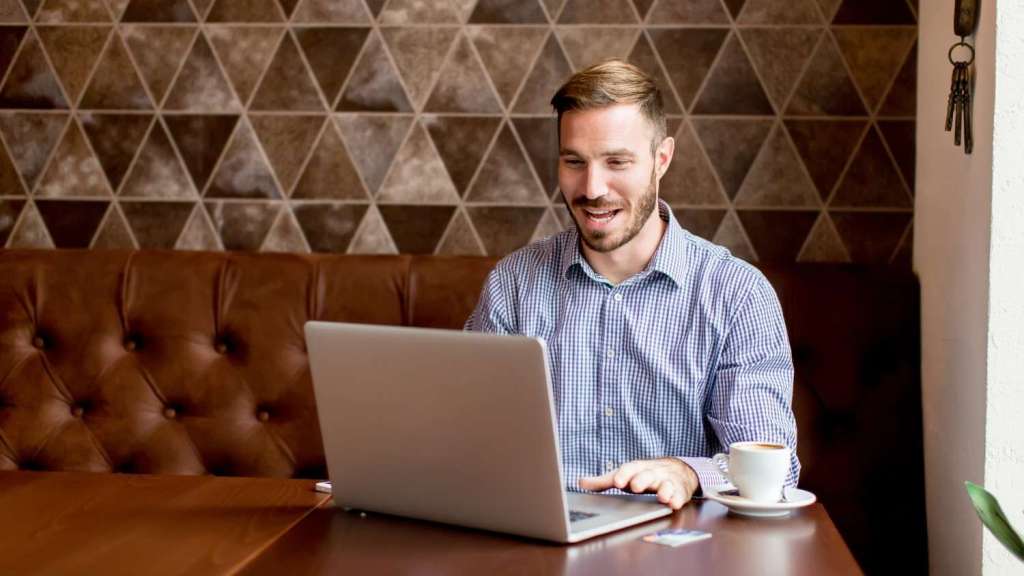 A young bearded male in a checked shirt sits at a table in a restaurant, drinking coffee and reading an article on his laptop about TransferGo vs Luminor Business