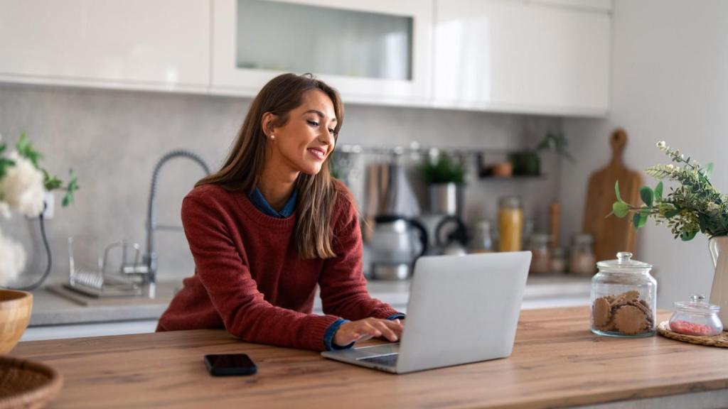 A young female smiles in her kitchen, whilst leaning on her kitchen top and reading an article on her laptop about 'TransferGo vs SEB Business'