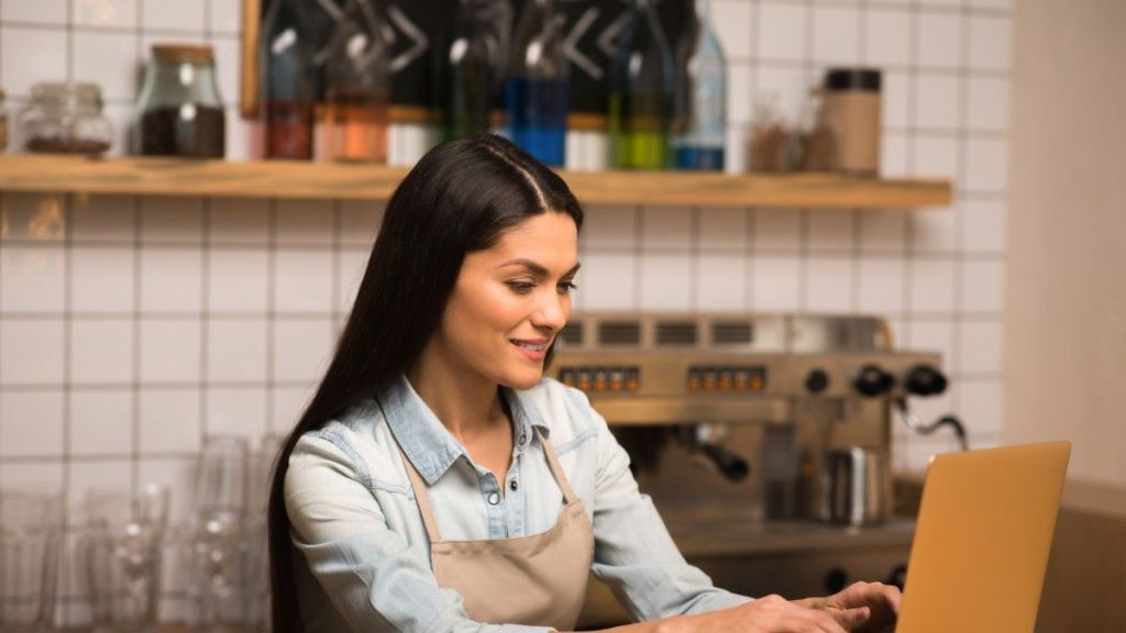 A young female with long dark brown hair wears a blue shirt and brown apron as she stands behind a cafe counter, types on her laptop and simplifies her supplier payments process