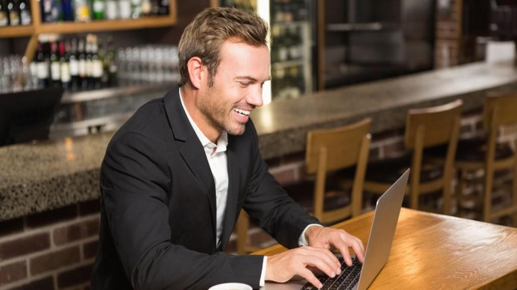 A handsome man in a black blazer and white shirt smiles as he sits down in a cafe and types on his laptop while simplifying his supplier payments process