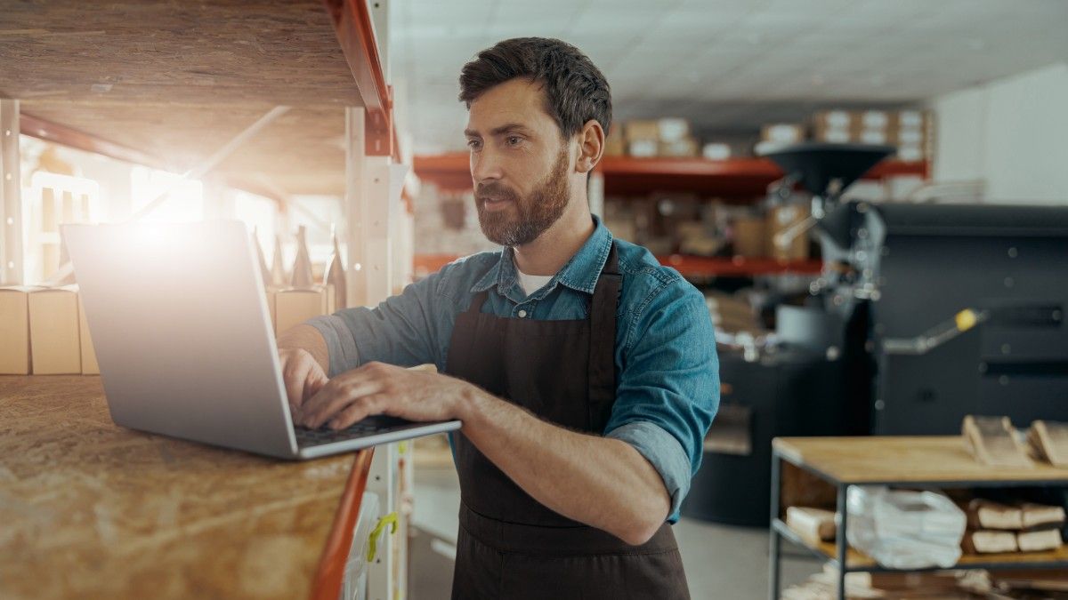 A young bearded man in a blue shirt and apron stands up in a cafe while typing on his laptop to simplify his supplier payments process