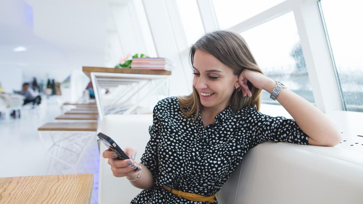 A young brunette female in a patterned shirt sits on the sofa and reads an article on her phone about how to pay international contractors