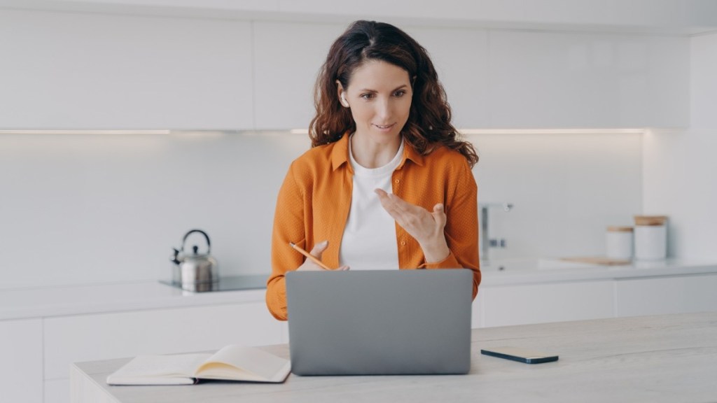 A young female with long brown hair wears an orange shirt and earphones as she stands in her white kitchen conversing in a meeting on her laptop about how to pay international contractors