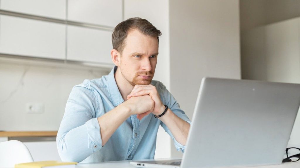 A young male in a light blue shirt sits at a desk and concentrates as he reads an article on his laptop about how to pay international contractors