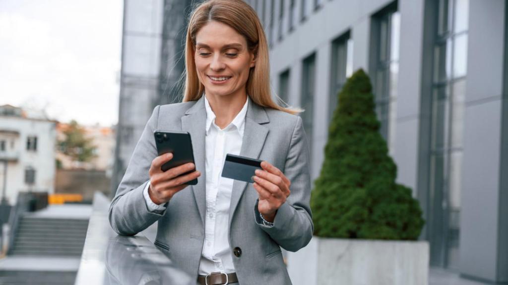 A young female in a grey blazer and white shirt smiles as she holds her phone and credit card, while reading an article titled 'Is Wise Business safe?'