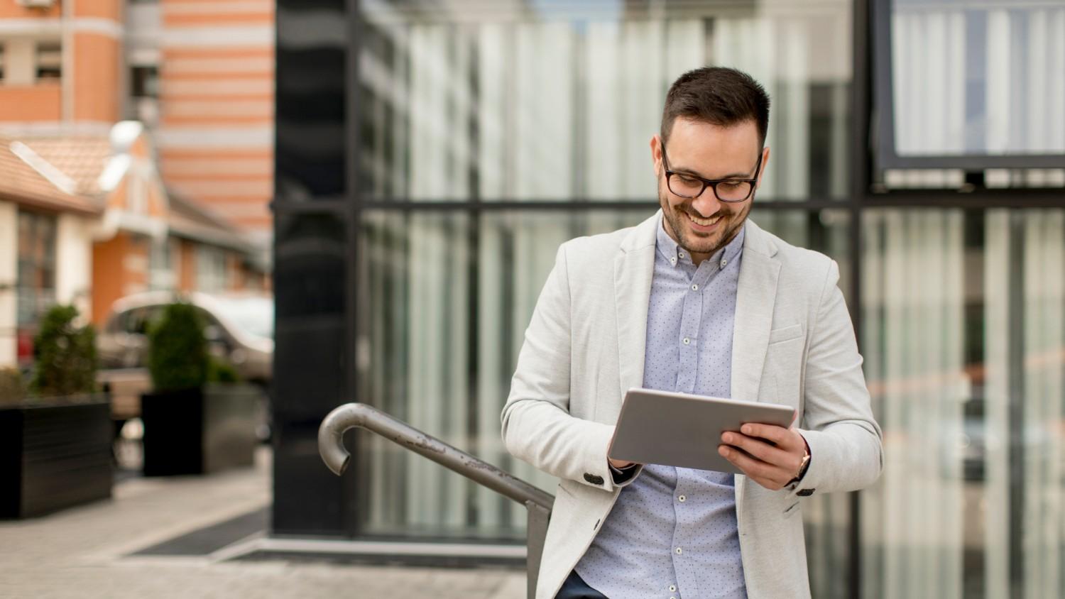 A young man in a casual shirt and suit stands outdoors and smiles as he holds his ipad and reads an article on 'Is Wise Business safe?'