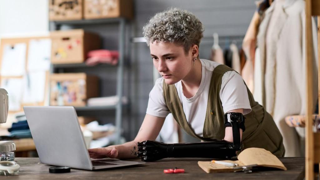 A young female with short curly hair and a bionic arm leans over her desk as she reads an article on her laptop about the best payment systems for small businesses