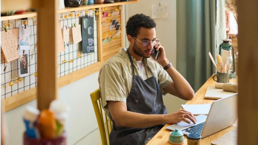 A young man in glasses and a T-shirt and apron sits at his desk as he talks on his phone and types on his laptop while researching the best payment systems for small businesses